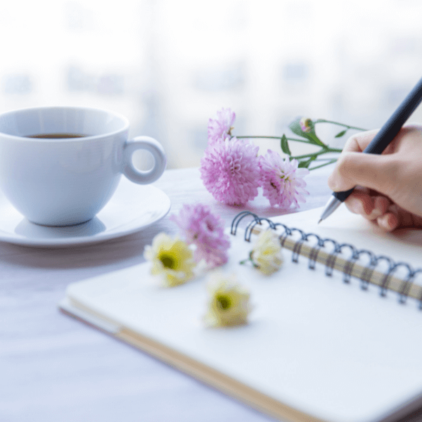 Frau schreibt in einem Block daneben steht eine Kaffeetasse und Blumen